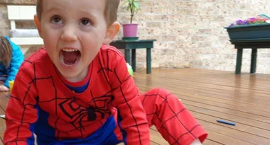 Child in Spider-Man costume, sitting on wooden floor, mouth open, playful expression. Indoor setting with potted plant behind.