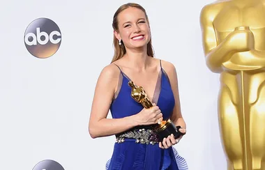 A woman in a blue dress smiles, holding an Oscar statue, standing next to a large gold Oscar statue.