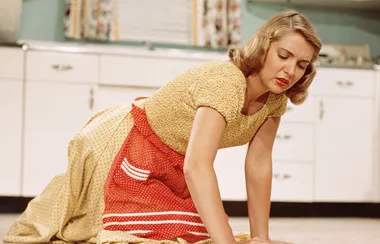 Woman in 1950s dress scrubbing floor, vintage kitchen in background.