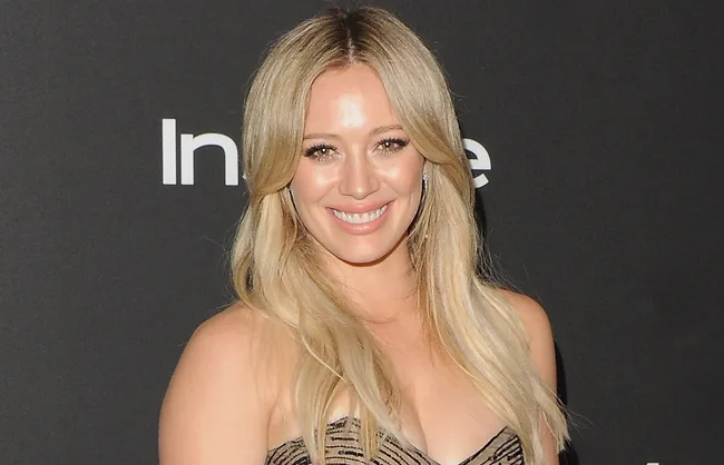 A person with long blonde hair smiling at an event with a dark background.