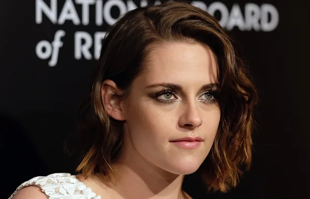Young woman with short, wavy hair in a white dress at an event with a "National Board of Review" backdrop.