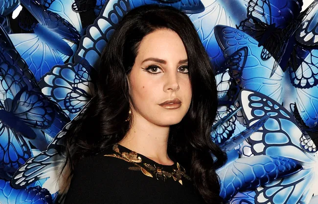 A person with long dark hair poses in front of blue butterfly art in a black outfit with gold details.
