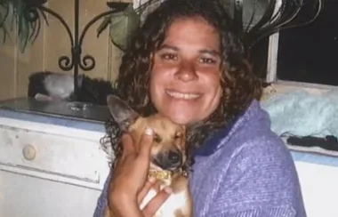 A person smiling and holding a small dog inside a room with plants and a window in the background.