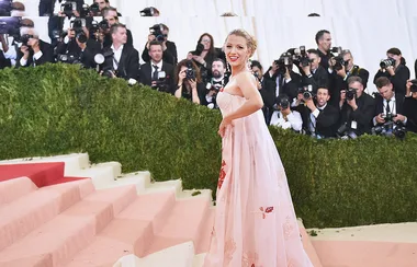 Woman in elegant pink dress on stairs, photographers in background at a glamorous event, likely a fashion gala.