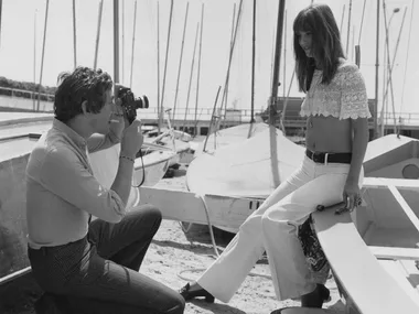 A man takes a photo of a woman in a lace top on a boat dock; sailboats are in the background.