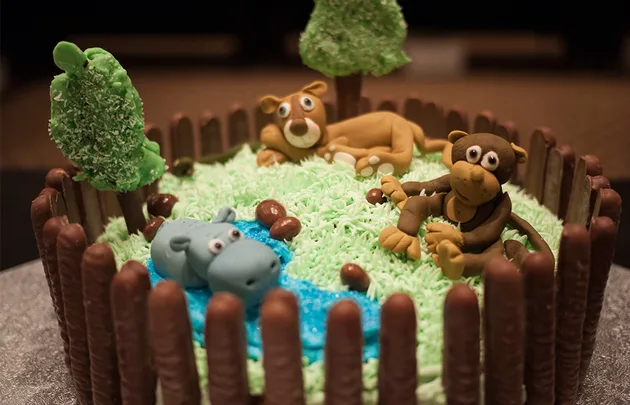 Cake with animal figurines: lion, hippo, and monkey in a grassy setting with chocolate fence and trees.