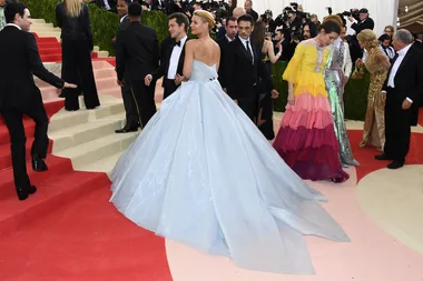 Woman in a flowing light blue gown at a formal event with others in colorful outfits on red carpet steps.