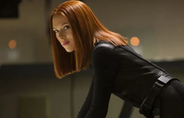A person with red hair in a black outfit leaning forward, possibly from an action film scene.