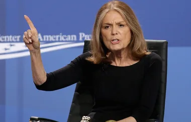 Lady speaking passionately at an event with a blue background and logos.