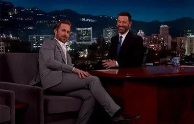 Two men in suits sitting and smiling on a talk show set with a cityscape background at night.