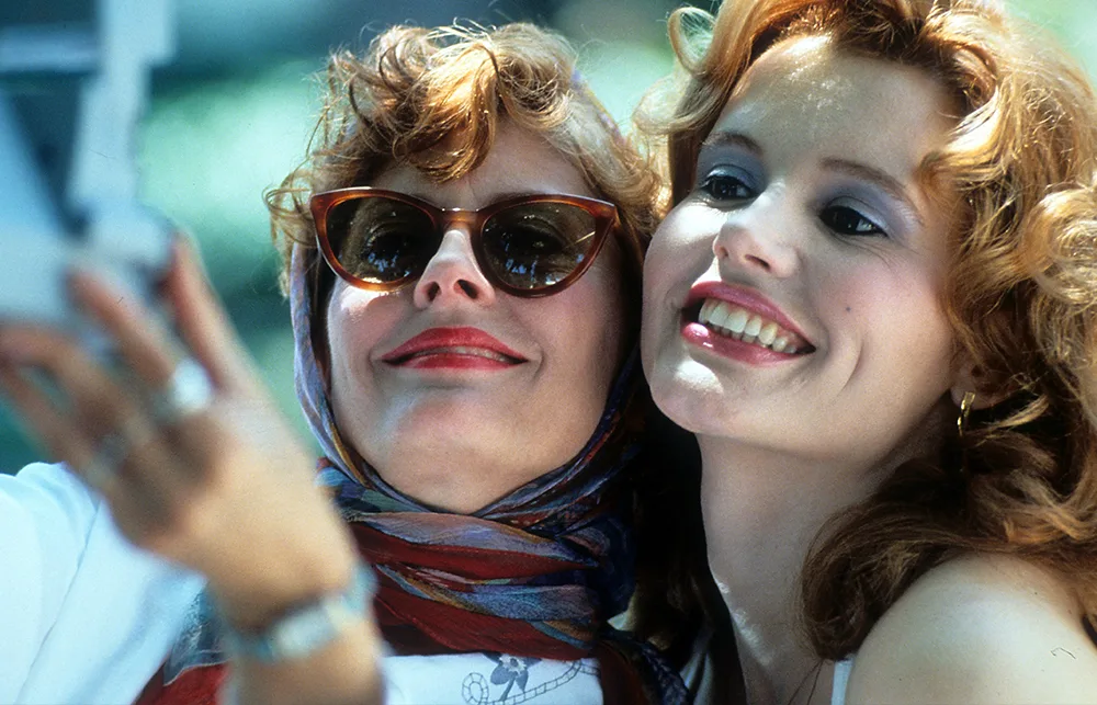 Two women smiling and taking a selfie, one wearing sunglasses and a scarf.