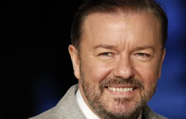 Smiling man with a beard, wearing a light grey suit jacket, against a dark background.