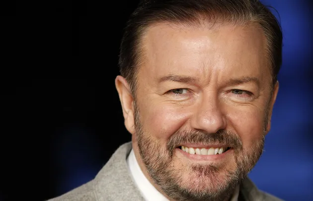 Smiling man with a beard, wearing a light grey suit jacket, against a dark background.