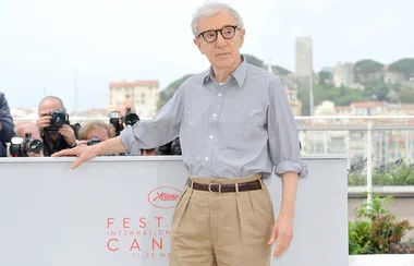Man in glasses standing at Cannes Film Festival, photographers in the background.