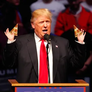 A man in a suit and red tie holds slices of pizza at a podium during a speech.