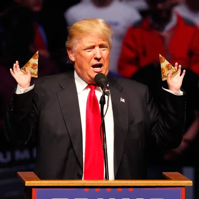A man in a suit and red tie holds slices of pizza at a podium during a speech.