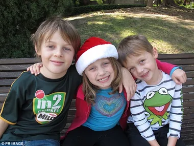 Three children sitting on a bench, girl in Santa hat, boys smiling, outdoor setting.