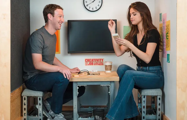 Two people sitting at a small table, one writing, the other laughing. Wall clock and motivational sign in background.
