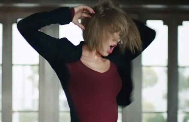A person mid-dance with tousled hair, wearing a maroon top and black jacket, in front of large windows.