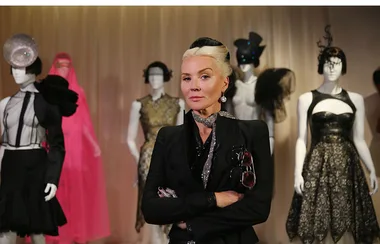 Blonde woman in black outfit stands confidently with arms crossed, surrounded by mannequins in elaborate fashion.