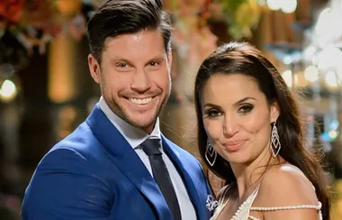 A couple posing elegantly, man in blue suit, woman in white dress with intricate earrings, smiling warmly.