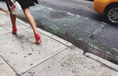 A person walks on a sidewalk wearing red high heels, carrying a shopping bag, with a yellow taxi passing by.