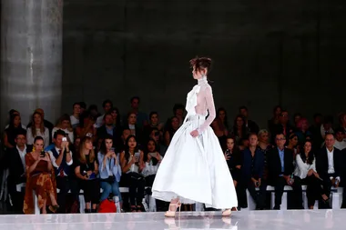 Model in a white dress walks runway, audience observes at fashion show.