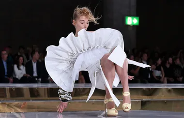 Model in white dress falls on runway, surrounded by seated audience.