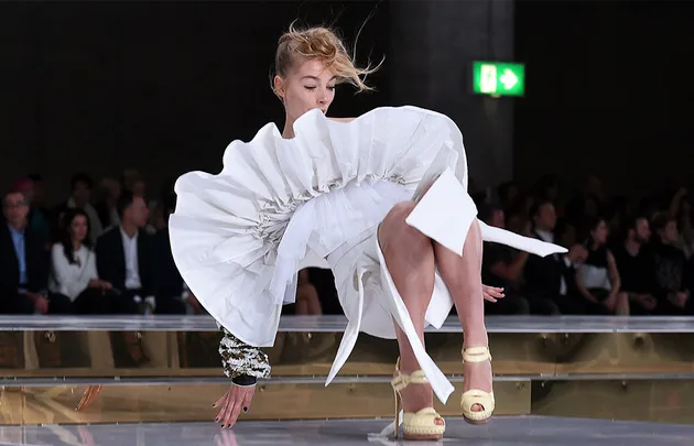 Model in white dress falls on runway, surrounded by seated audience.
