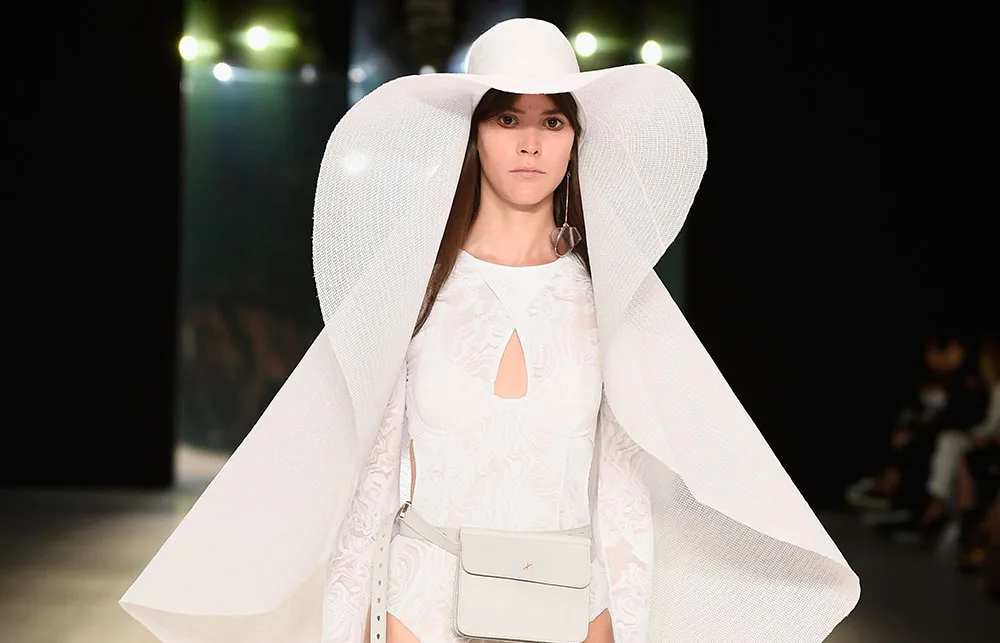 A model in a large white hat and dress walks the runway, wearing a white belt bag.