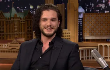 A person with curly hair in a suit smiling on a talk show set with a cityscape backdrop.