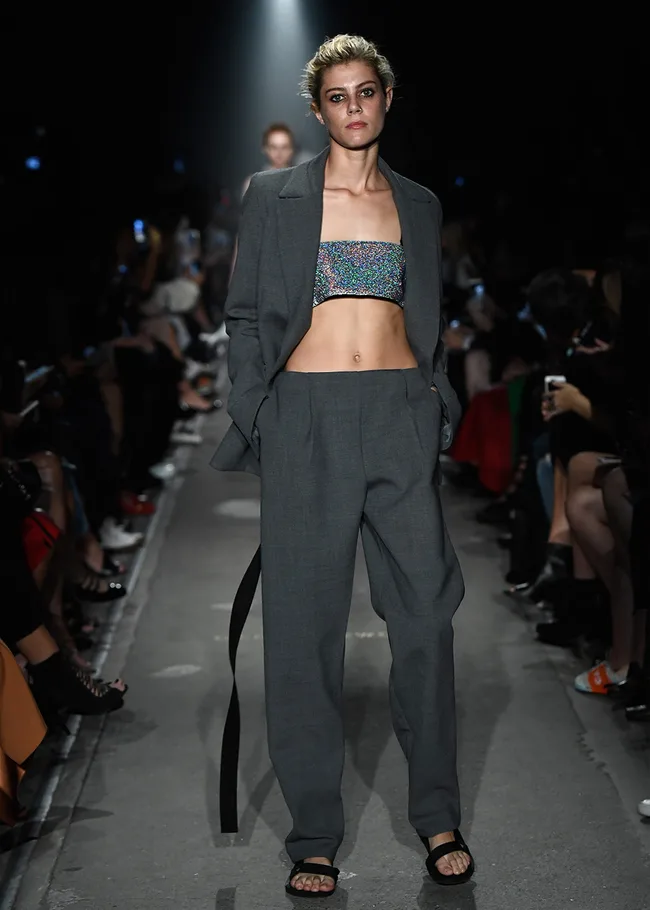 Model in gray suit with a colorful crop top walks on a dimly lit runway.