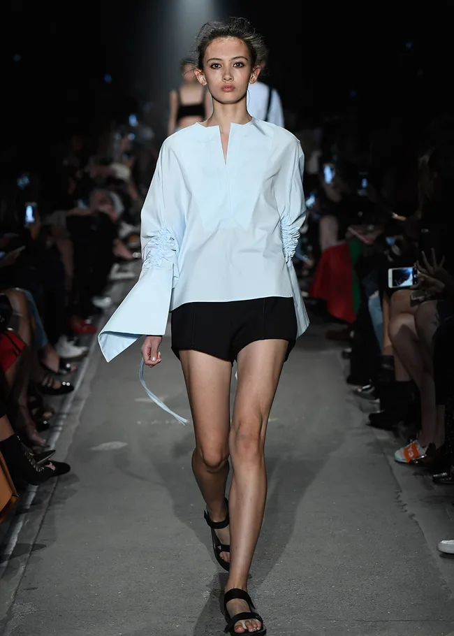 Model walking on a runway wearing an oversized light blue shirt and black shorts with an audience capturing the scene.