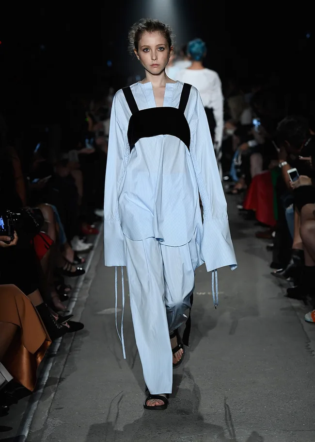 Model on a runway wearing a loose-fitting, long-sleeve blue outfit with black straps, surrounded by audience.