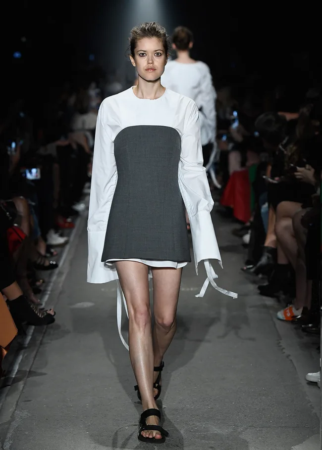 Model walks runway in monochrome dress with long sleeves, black sandals, and hair up, during fashion show.