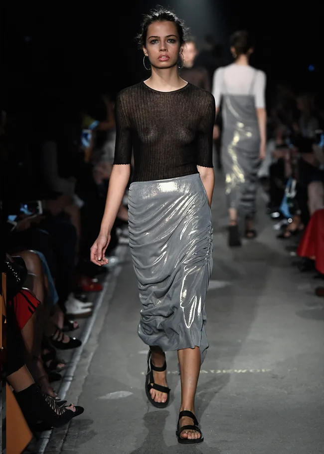 Model in sheer black top and metallic silver skirt walking on a runway during a fashion show.