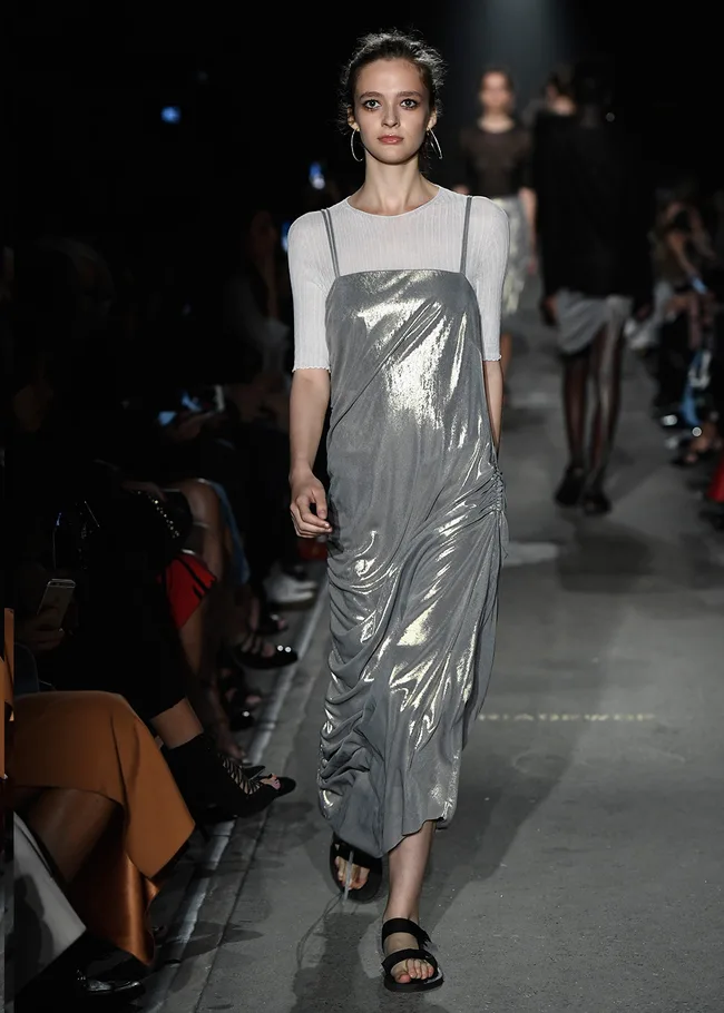 Model in metallic silver dress and white top walks runway during a fashion show, audience in background.