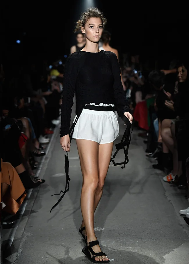 Model walks runway in black long-sleeve top and white shorts during a fashion show, with audience seated on both sides.