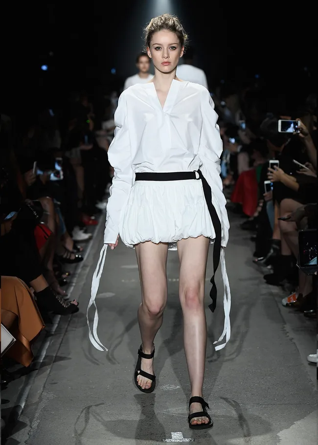 Model in a white outfit walks the runway at a fashion show, surrounded by an audience taking photos.