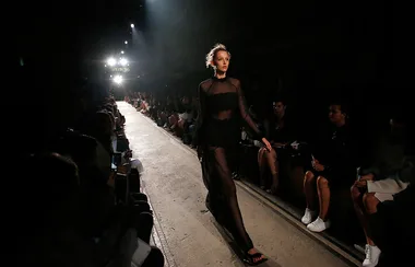 Model walks down a fashion runway in a sheer black dress, with audience seated on both sides.