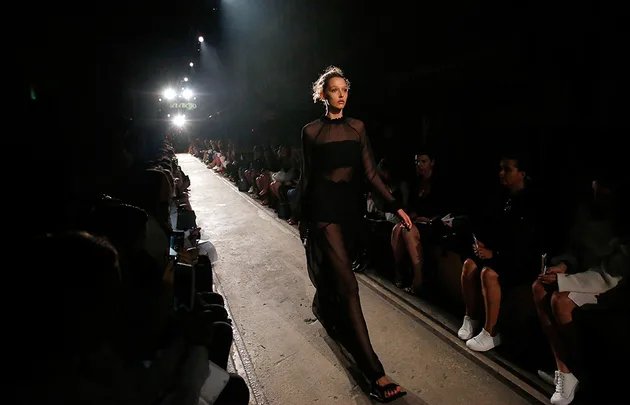 Model walks down a fashion runway in a sheer black dress, with audience seated on both sides.