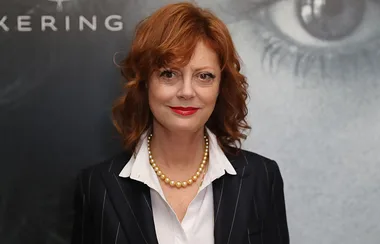 A person with red hair and a black suit poses, wearing a white shirt and pearl necklace, with a blurred backdrop behind.