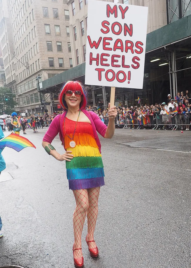 Person in colorful dress at pride parade holds sign: "My Son Wears Heels Too!"