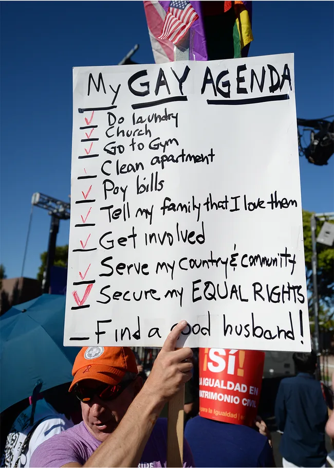 Man holds sign titled "My GAY AGENDA" with a humorous to-do list at a protest, featuring a small US flag on top.
