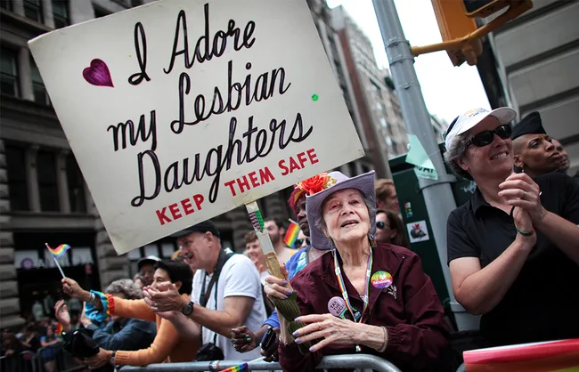 Elderly woman in a hat holds a sign reading "I adore my lesbian daughters, keep them safe" at a Pride parade.