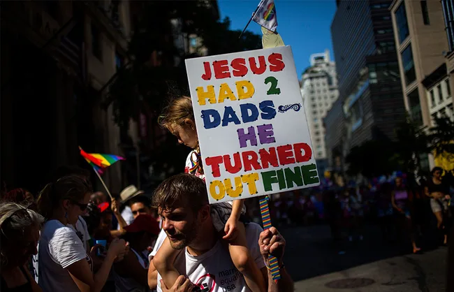 Man carries child holding sign reading "Jesus had 2 dads he turned out fine" at a pride parade, with a rainbow flag nearby.