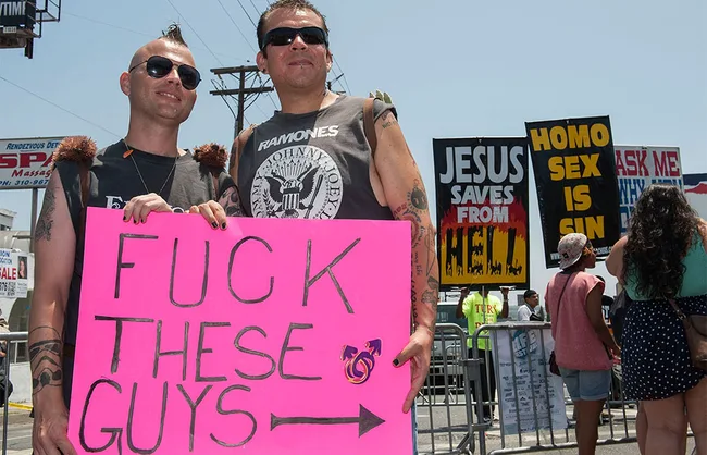 Two men wearing sunglasses hold a pink sign with profanity, countering anti-gay protestors' signs in the background.