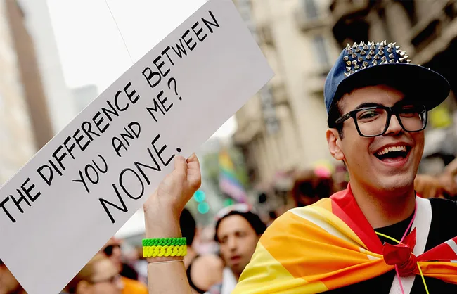 Young person at a pride event, wearing a spiked cap and rainbow flag, holds a sign reading "The difference between you and me? None."