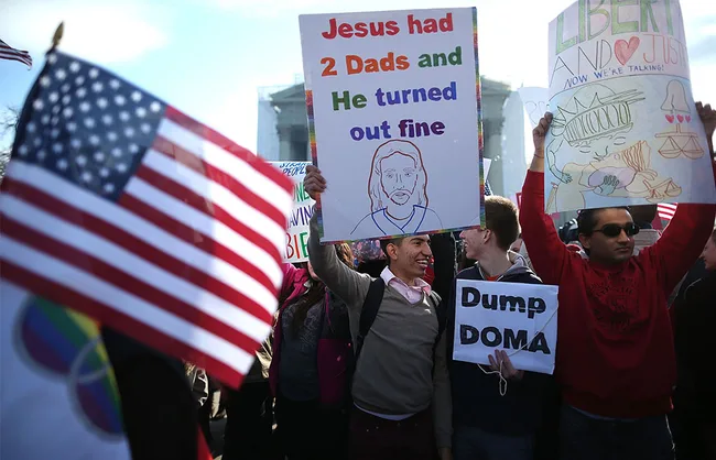 Crowd at a protest holding signs, one reads "Jesus had 2 Dads and He turned out fine," and another "Dump DOMA," with U.S. flags.