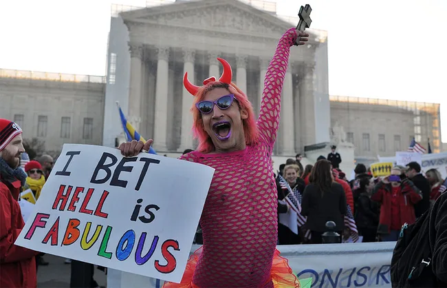 Person in pink outfit and devil horns holds a sign reading "I bet hell is fabulous" in a crowd at a protest or event.